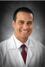Professional headshot of Rodrigo Antonio Figueroa, MD, wearing a white lab coat and red patterned tie against a dark studio background.