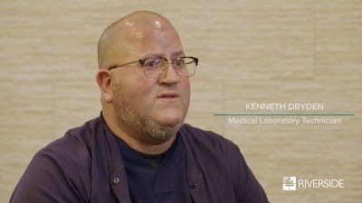 Bald Caucasian Male with glasses and a purple shirt sitting in front of a tan wall