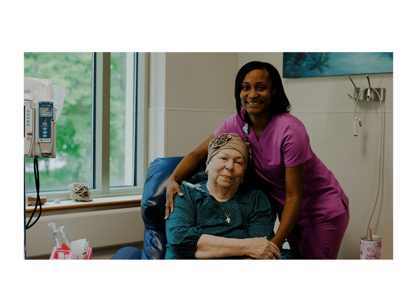 African American woman in pink scrubs with her arm around a Caucasian woman in a chair with a scarf on her head