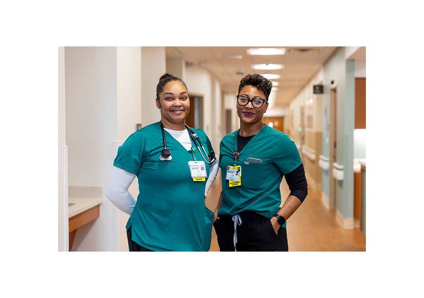 Two healthcare team members wearing scrubs standing together in a hospital hallway.