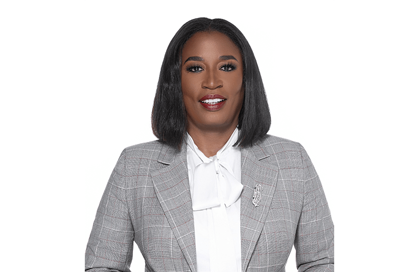 A smiling woman with shoulder-length black hair wears a gray plaid blazer and white blouse, facing the camera in a polished professional studio portrait.