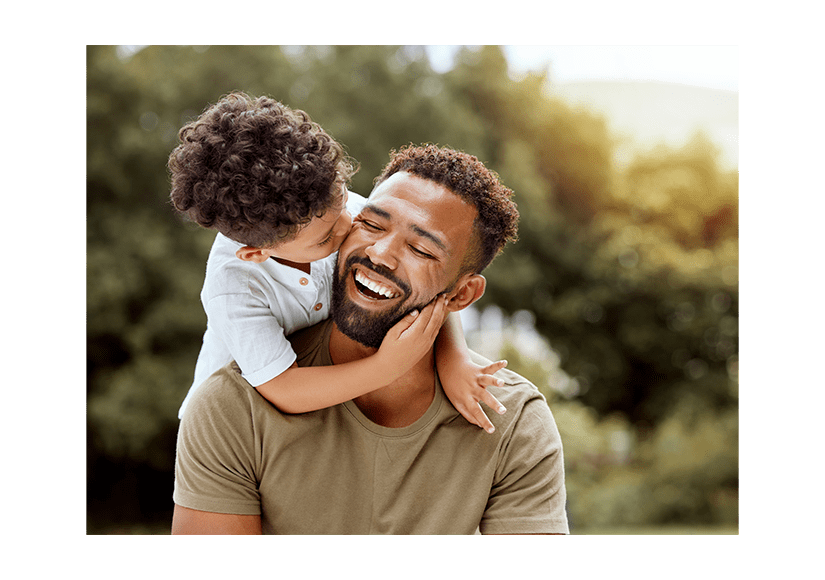 A joyful Black father carries his young child outdoors as the child hugs and kisses his cheek, with warm sunlight and greenery creating a loving scene.