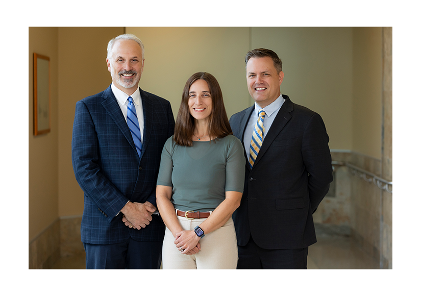 Three smiling professionals—two men in suits and a woman in a teal top—pose together in a bright hallway, conveying warmth and teamwork.