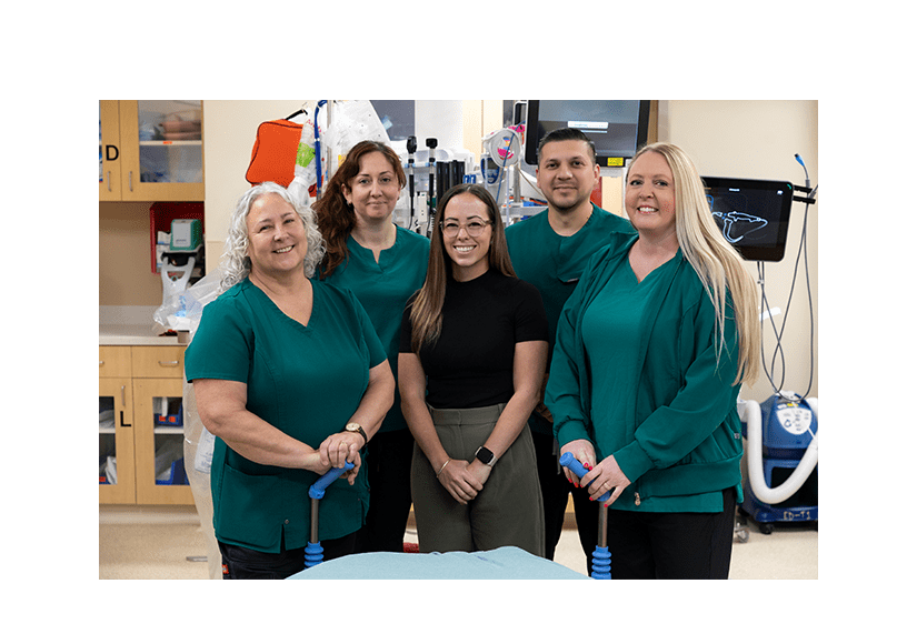 Five smiling healthcare professionals pose in a hospital room, wearing teal scrubs and holding blue medical tools, conveying teamwork.