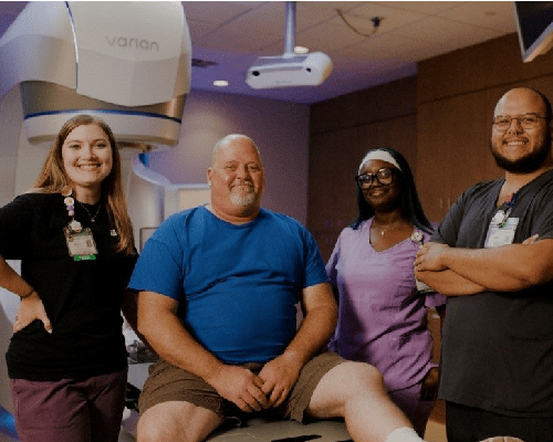 Riverside Health patient with clinical care team in radiation therapy treatment room, highlighting compassionate care, teamwork and advanced medical technology.
