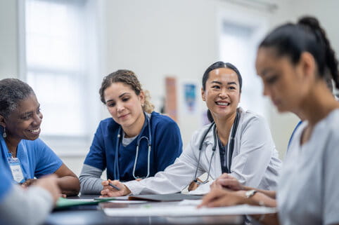Ready to Apply? Take the Next Step A small group of doctors and nurses gather around a meeting table to discuss patient cases. They are each dressed professionally and have various, files, notes and papers scattered between them as they reference them throughout the meeting.