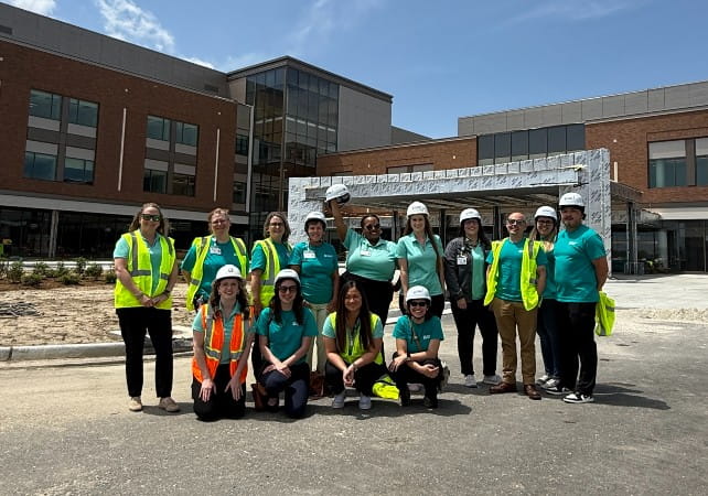 A group of construction professionals wearing hard hats and high‑visibility vests pose in front of a modern building under construction, featuring large glass windows and a metal framework.
