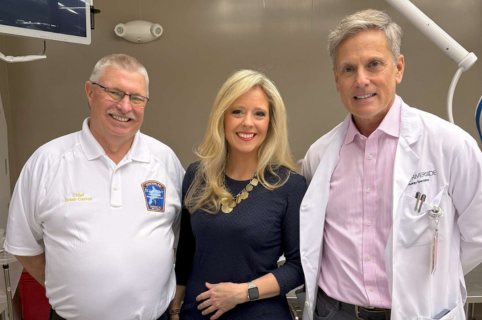 Three adults stand together in a clinical room, with medical equipment visible in the background.