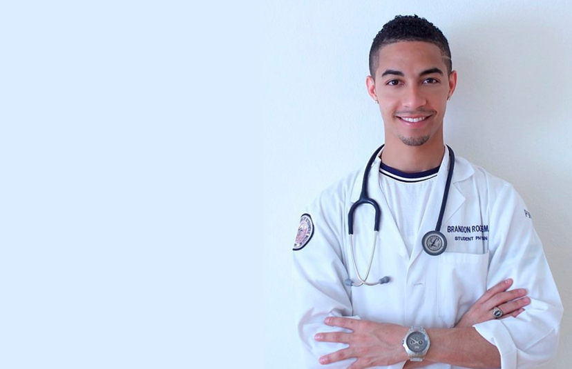 Healthcare student in a white coat with stethoscope standing against a light wall.
