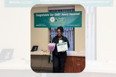  A healthcare employee stands indoors holding a DAISY Award certificate and a bouquet of flowers beneath a congratulatory DAISY Award banner.
