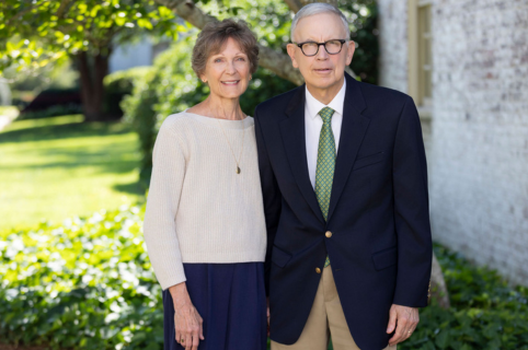 Two adults standing side by side outdoors in front of a light-colored building and landscaped greenery. One person wears a cream sweater and dark skirt, and the other wears a navy blazer, green patterned tie, and khaki pants.