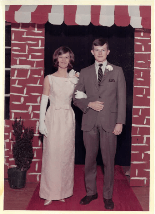 Vintage photograph of two young adults dressed for a formal event, standing together arm in arm, reflecting a cherished moment from earlier years.