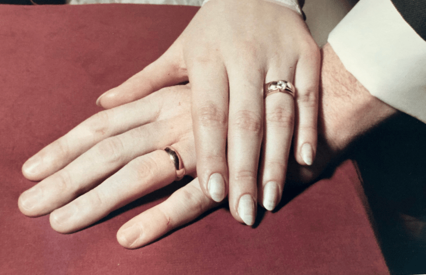 Close-up of two hands wearing wedding rings resting together on a burgundy surface.