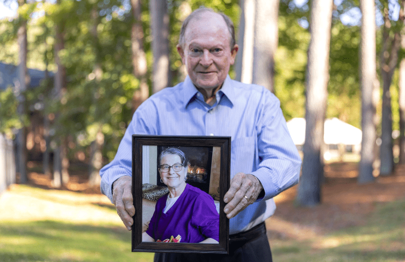 Person standing in a peaceful wooded setting holding a framed photograph of a loved one in remembrance, highlighting a personal story of hospice care and legacy.