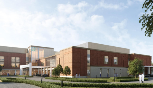 A modern hospital exterior with red brick walls, large windows, and a glass entrance. People in white coats walk nearby. The sky is bright and partially cloudy.