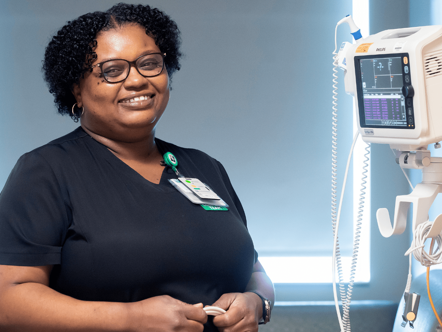 Smiling nurse in glasses and scrubs stands beside medical equipment with a monitor displaying vital signs. The setting is professional and caring.