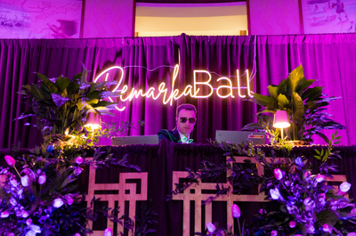 DJ in sunglasses at a vibrant event with lush plants, purple lighting, and a neon "RemarkaBall" sign, creates an energetic and festive atmosphere.