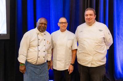 Three chefs wearing white uniforms stand smiling against a blue curtain backdrop. The mood is professional and celebratory.