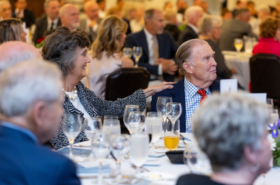 A lively banquet scene with a woman smiling and resting her hand on a man's shoulder. Both are seated among elegantly set tables and attentive guests.