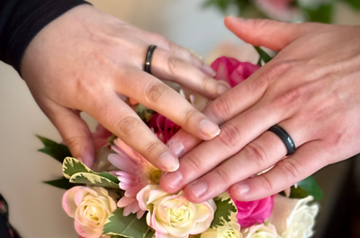 Two hands with black rings placed on a pink and white flower bouquet, symbolizing unity and love. The scene conveys a romantic and joyful tone.
