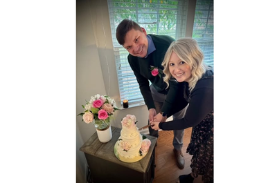A smiling couple cuts a three-tiered white cake adorned with pink flowers. A vase of pink and white roses is nearby. The setting is bright and joyful.