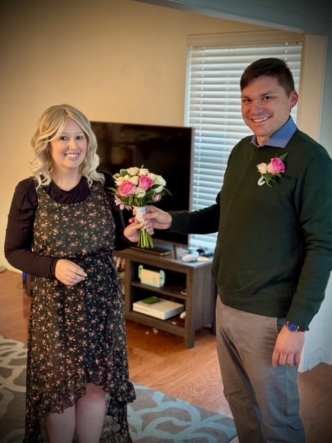 A smiling woman in a floral dress receives a bouquet of flowers from a man in a sweater with a matching boutonniere in a cozy living room.