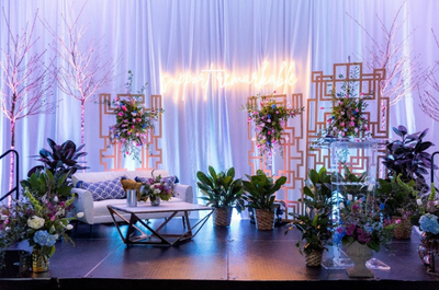 Elegant stage with white drapes, glowing neon sign, floral arrangements, and greenery. Cozy white couch and table set a romantic tone.