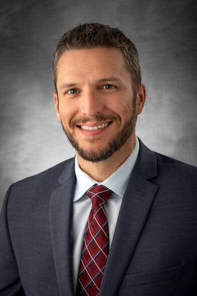 A smiling man in a suit with a red and blue tie against a gray background. The tone is professional and approachable.