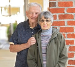 Caucasian Older Man in a dark blue shirt stands next to an older Caucasian woman in a green jacket in front of a brick wall