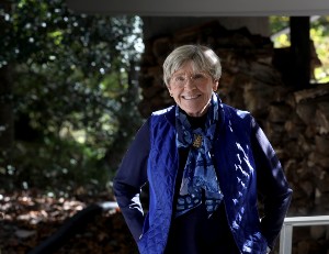 Older woman standing outside in front of a tree with a blue shirt smiling