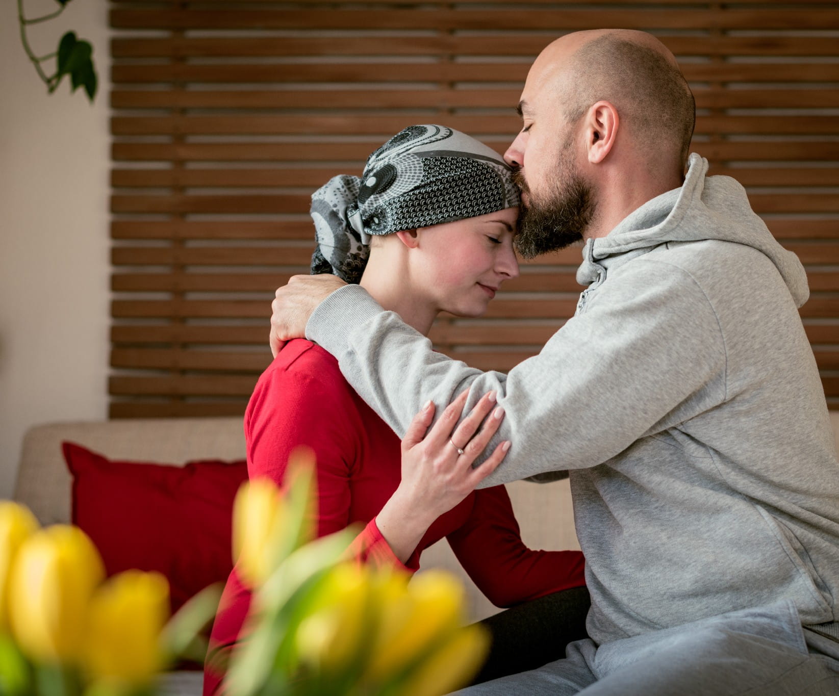 A man gently kisses a woman's forehead as they embrace on a sofa, conveying support and care. She wears a headscarf, and a vase of yellow flowers is in the foreground.