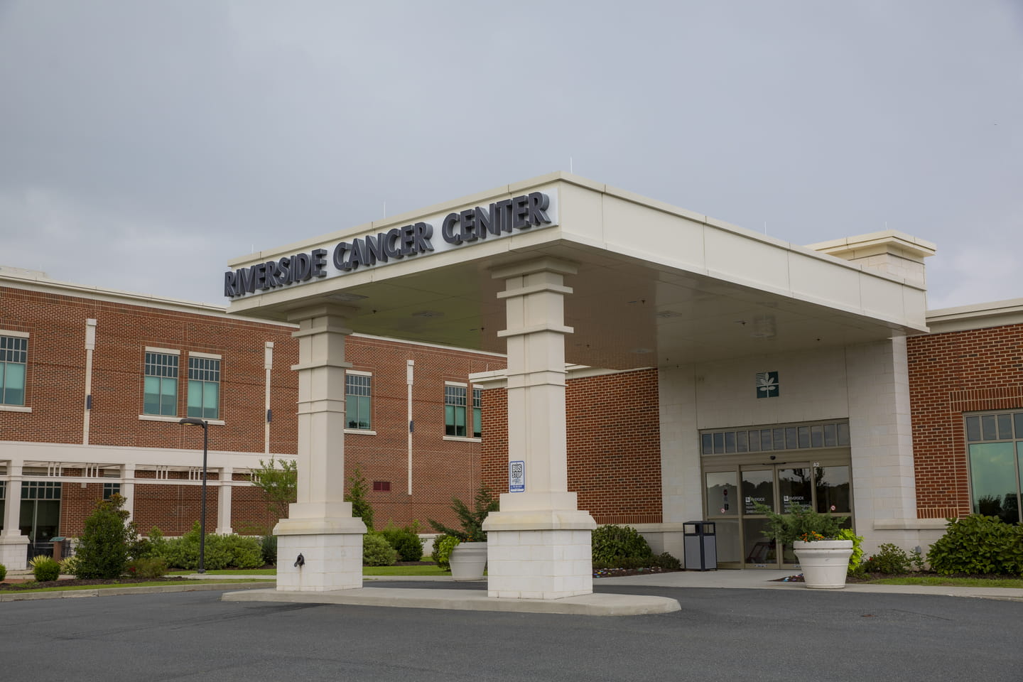 Entrance of Riverside Oncology Specialists Shore with a large white canopy and sign, surrounded by green shrubs against a brick building under a cloudy sky.