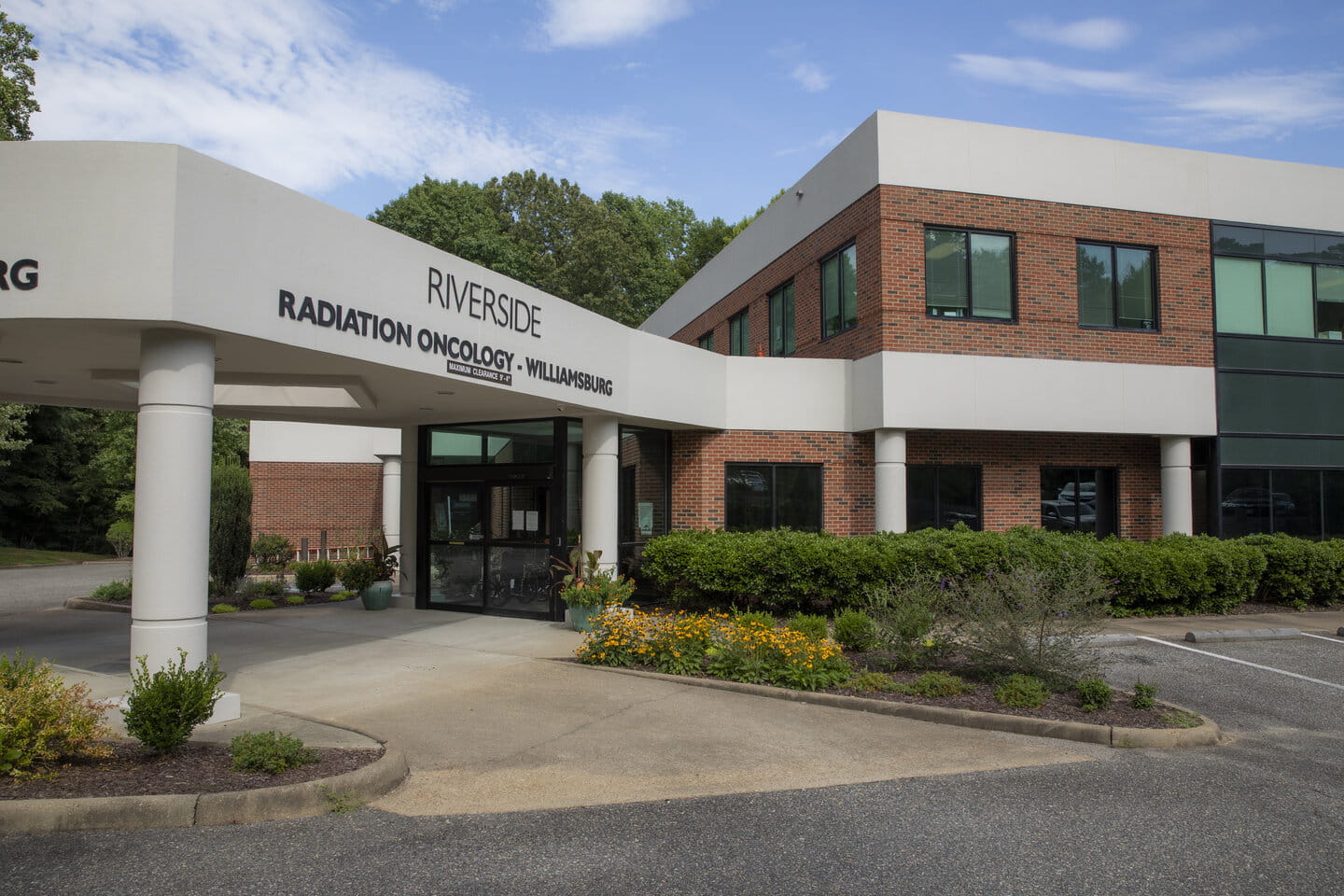 Modern brick medical building with "Riverside Radiation Oncology - Williamsburg" sign, surrounded by greenery and colorful flowers under a blue sky.