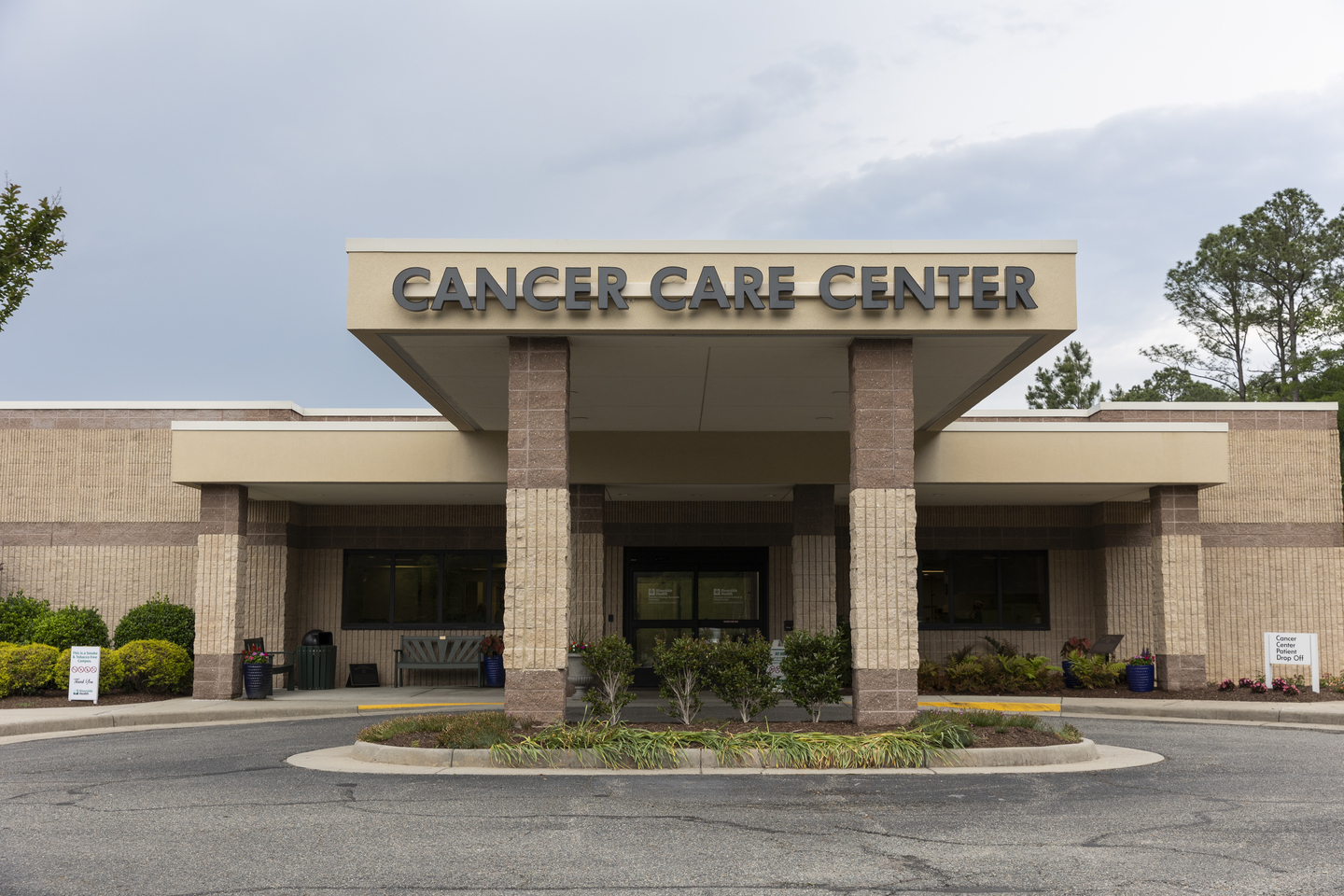 Exterior of Radiation Oncology Specialists Gloucester with a beige brick facade and sign. The entrance is flanked by well-maintained shrubs, under a clouded sky, creating a calm, welcoming atmosphere.