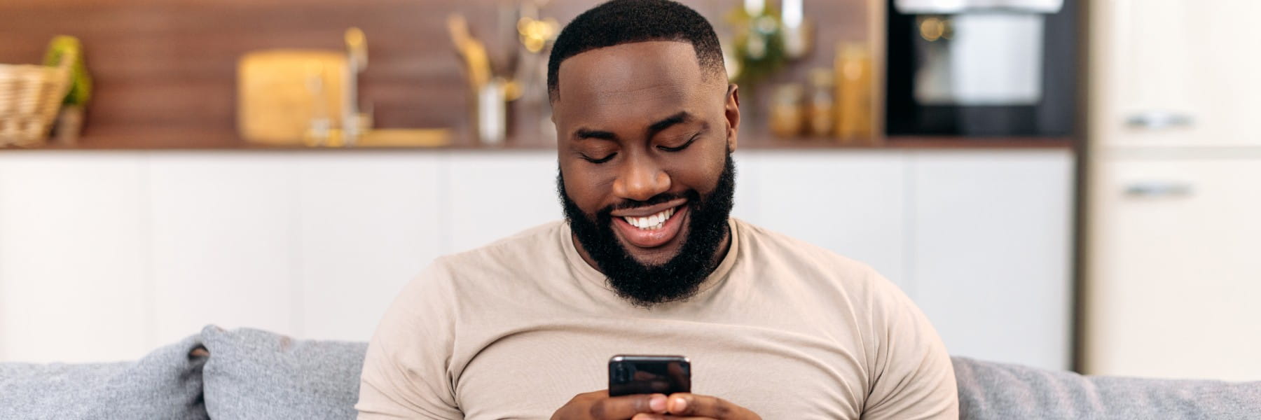 Modern attractive positive african american man in casual wear, sits at home on sofa, using his smartphone, 
