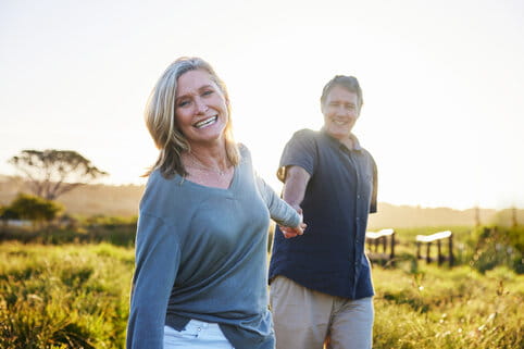 A joyful couple holds hands, walking in a sunlit field. The woman smiles warmly at the camera, evoking happiness and connection in a serene landscape.