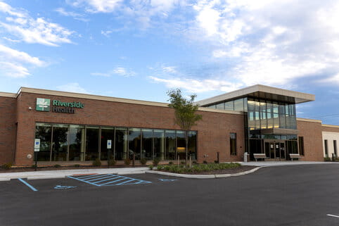Modern healthcare building with brick facade, large glass windows, and a blue sign reading "Riverside Health." Clear sky and accessible parking visible.