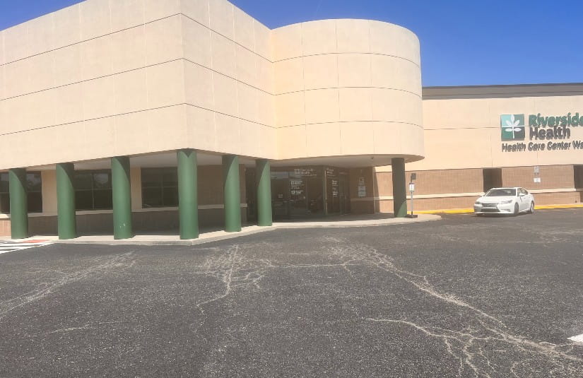 Large beige building with green columns, labeled "Riverside Health Care Center West." A white car is parked nearby under a clear blue sky.