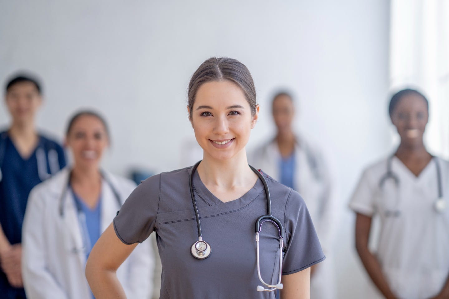 Group of healthcare providers standing together in a bright medical clinic, including doctors and nurses in scrubs and white coats, representing a professional care team.