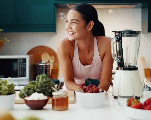 Person leaning on a kitchen counter with fresh fruits and vegetables, with a blender and modern kitchen appliances in the background
