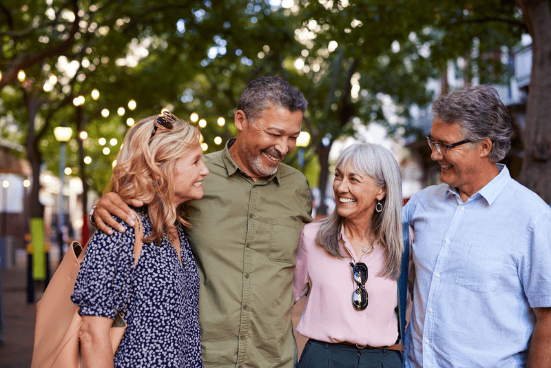 Four older friends sharing a laugh while walking outside. Happy multiethnic seniors couples enjoying a walk and smiling at each other during spring vacation in european city. Group of retired elderly men and cheerful women spending time together during trip.