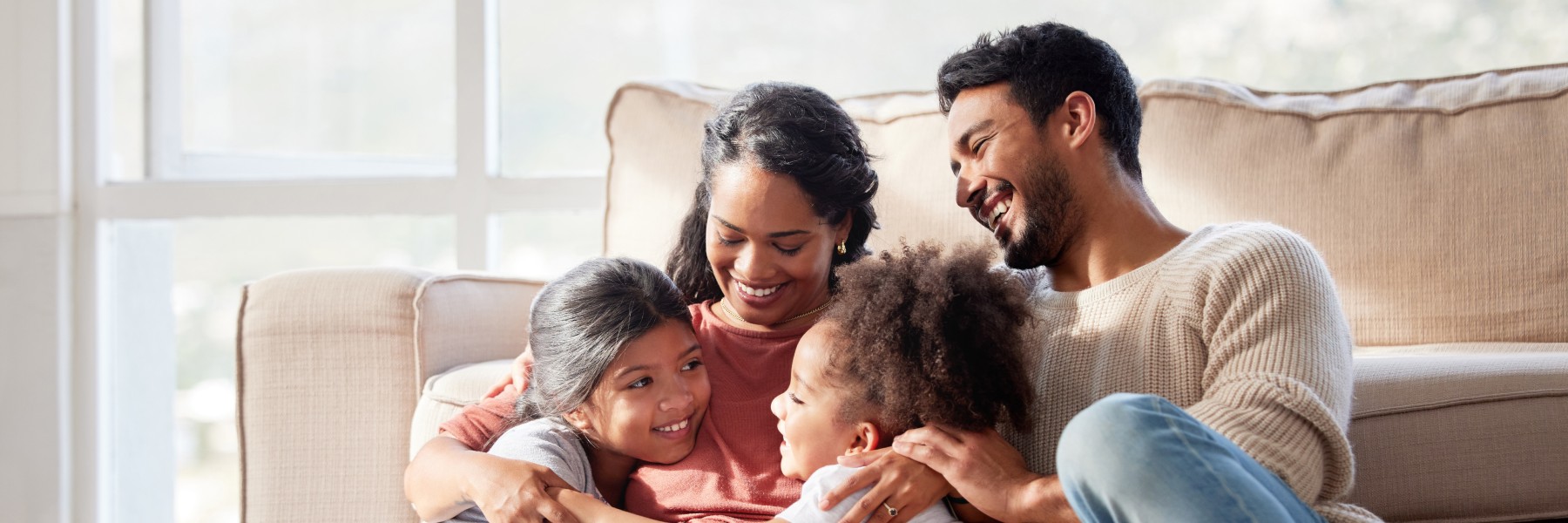 A happy family of four sits on a couch in a sunlit room. The parents embrace their two young children, smiling warmly, creating a joyful and loving atmosphere.