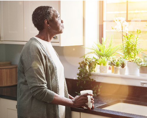 A thoughtful senior woman having a cup of coffee in the kitchen and looking out of her window at home. Mature woman drinking tea and thinking 