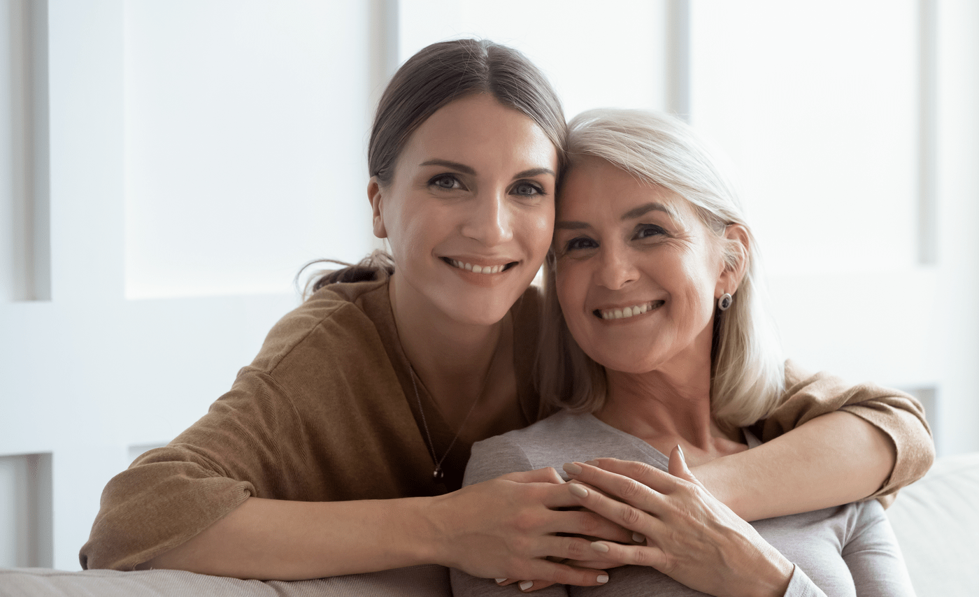 Head shot close up portrait smiling young woman embracing from back sitting on couch middle aged mother. Happy two generations retired mom and grown up daughter posing for photo in living room.