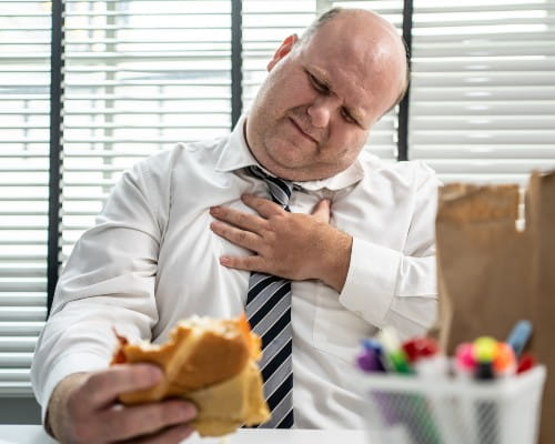 Guy suffering from heartburn sitting on chair and holding hamburger wearing shirt .Acid reflux, pain in stomach concept.