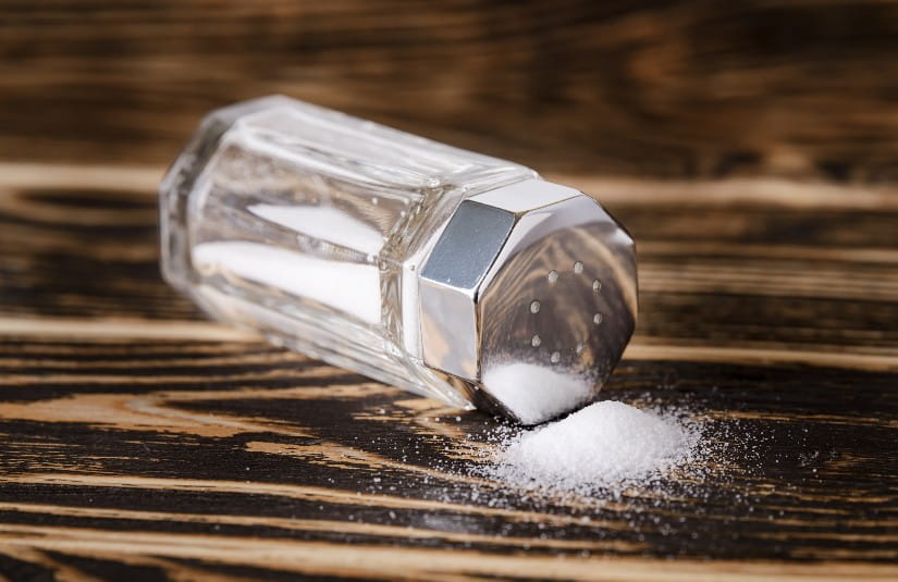 Some salt spilled out of the glass salt shaker with a silver metal lid lies on its side on a dark wooden table