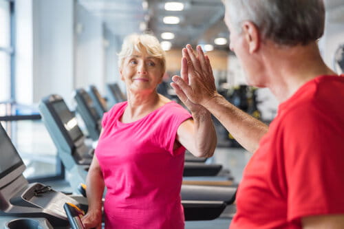Senior man and woman walking on treadmills and giving each other high five at gym. Senior couple enjoying at rehabilitation centre.