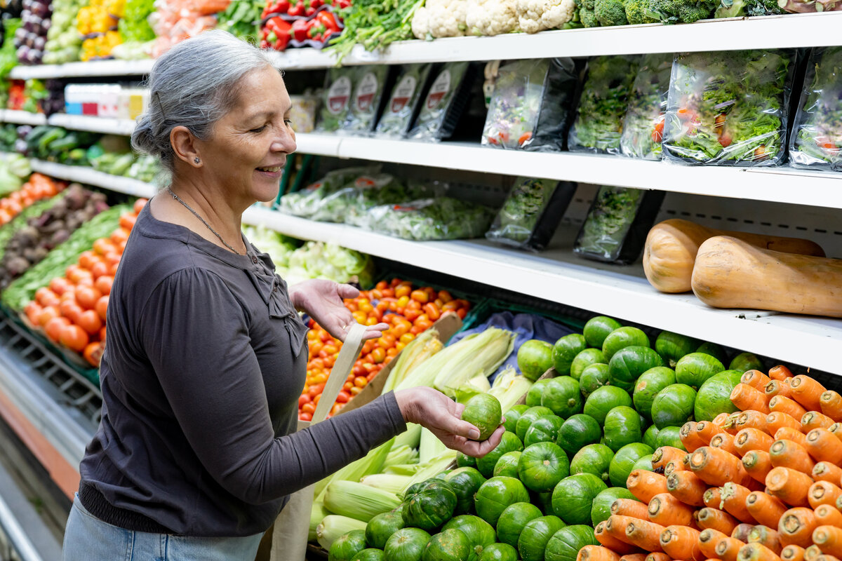 Cheerful senior woman at the supermarket choosing vegetables while holding a reusable bag -