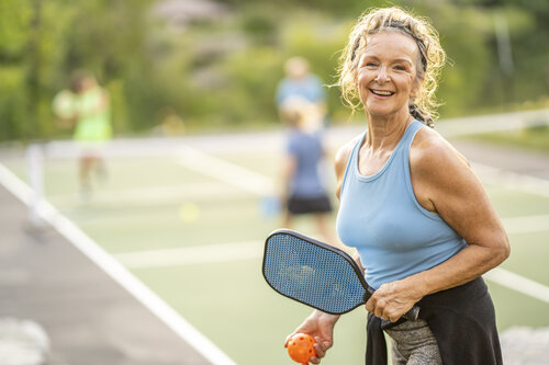 A middle aged woman poses for a portrait while on a Pickleball court. She is dressed comfortably in athletic wear and is holding a racquet and ball as she smiles for the camera.