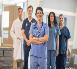 A multi-ethnic group of nurses and a doctor are standing together in their hospital clinic. They are all smiling and looking at the camera.